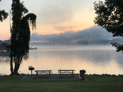 A serene lakeside view at dawn, featuring a picnic area, mist over the water, and soft clouds in the sky.