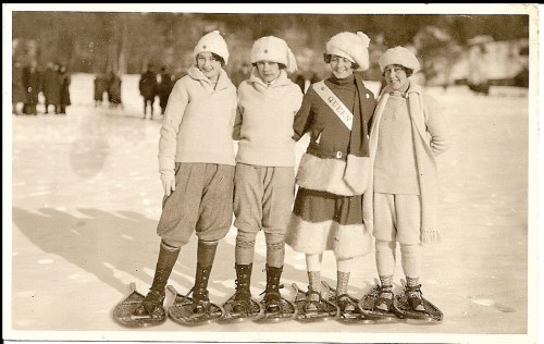 Four girls in vintage winter attire stand on snowshoes, smiling on a snowy landscape, with a crowd in the background.
