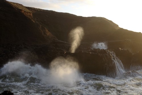 Waves crash against rocky cliffs, creating mist and spray under a golden sunset sky.