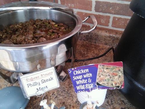 A buffet setup featuring Philly steak meat in a chafing dish and a sign for chicken soup with white and wild rice.