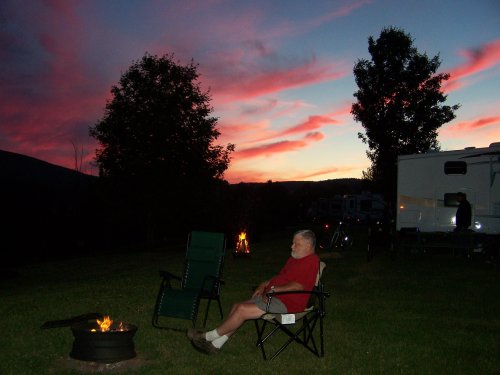 A person sits by a campfire at dusk, with colorful clouds and a camper in the background.