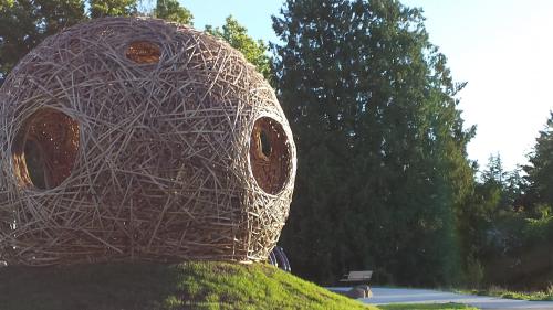 A large, spherical sculpture made of intertwined branches, set in a green park with trees in the background.