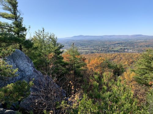 Scenic view from a rocky overlook, showcasing colorful autumn foliage and distant mountains under a clear blue sky.