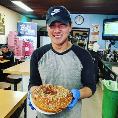 A smiling person in a cap holds a large, colorful donut with sprinkles in a cozy donut shop.
