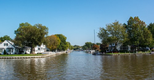 A serene waterway lined with trees and houses under a clear blue sky, reflecting the calm water.