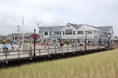 A boardwalk scene with shops, people walking, and a view of the beach grass under a cloudy sky.