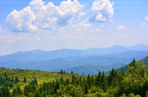 A scenic view of rolling mountains and lush green forests under a blue sky with fluffy white clouds.
