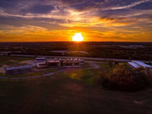 Aerial view of a landscape at sunset, featuring a building and open fields under a colorful sky.