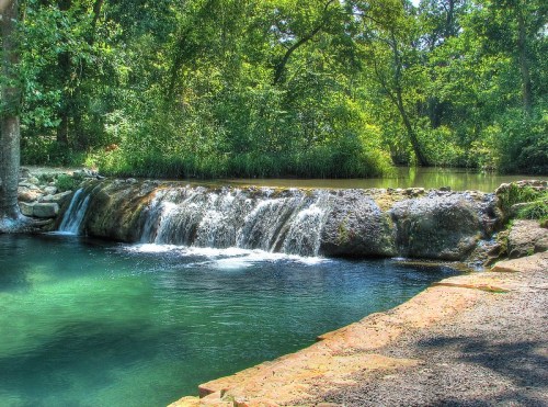 A serene waterfall cascades over rocks into a clear, tranquil pool surrounded by lush greenery.