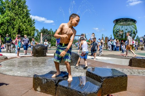 A young boy plays in a splash pad, surrounded by other children and families enjoying a sunny day at the park.