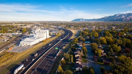Aerial view of a highway with traffic, industrial buildings, and residential areas surrounded by mountains and trees.