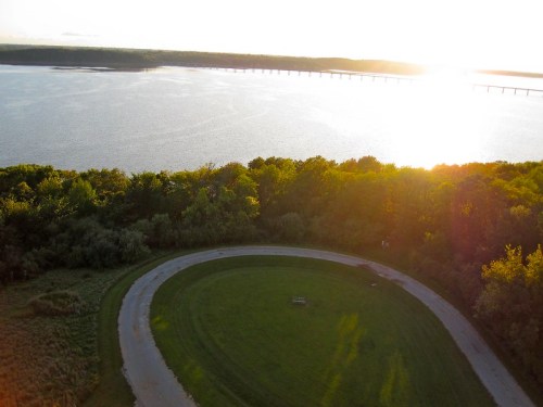 Aerial view of a curved path through greenery, overlooking a river and a bridge at sunset.