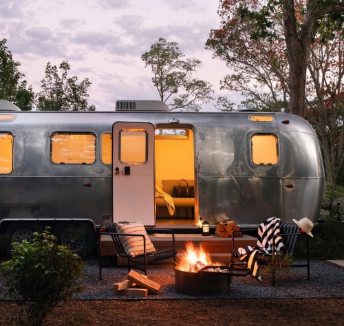 A cozy Airstream trailer with an open door, surrounded by a fire pit and outdoor seating, set in a serene landscape.