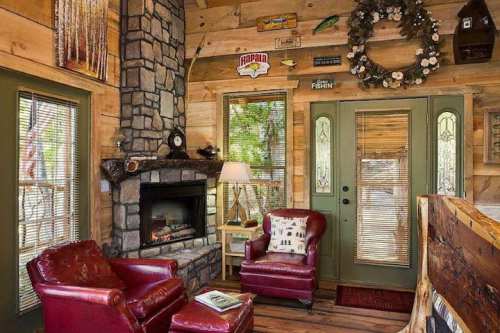 Cozy cabin interior featuring a stone fireplace, red armchairs, wooden walls, and decorative accents.
