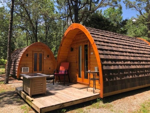 Two wooden cabins with curved roofs, surrounded by trees, featuring a small porch and outdoor seating.