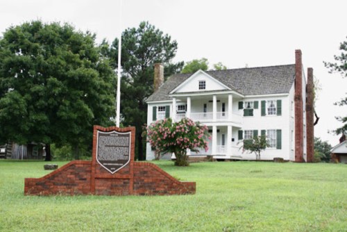 Historic white house with green shutters, surrounded by trees and a plaque in front on a grassy area.