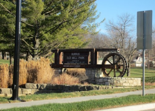 Sign for Burrus Old Mill Park featuring a wooden cart wheel, surrounded by grass and trees.