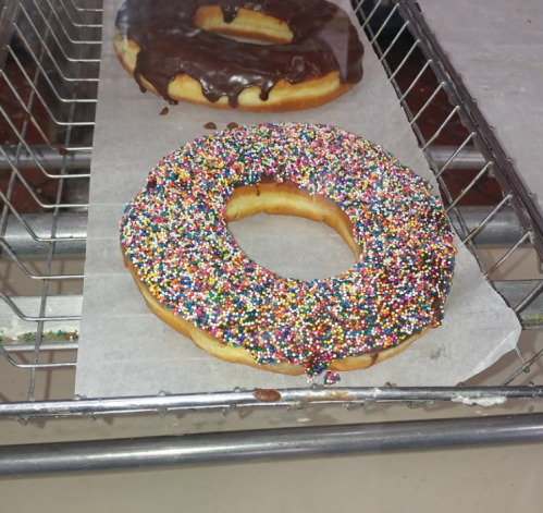 A colorful donut covered in sprinkles sits on parchment paper, with a chocolate-glazed donut in the background.