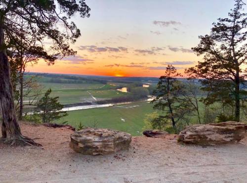 A scenic view of a sunset over a river and green fields, framed by trees and rocky terrain.