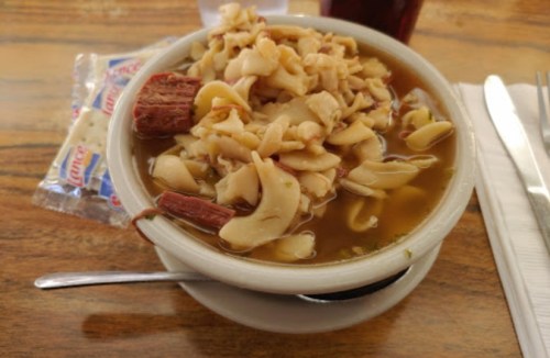 A bowl of soup with noodles and chunks of meat, served with a spoon and a side of crackers on a wooden table.