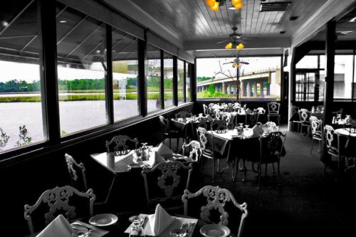 A dining area with elegant tables and chairs, large windows overlooking a river and bridge, in a partially black-and-white setting.