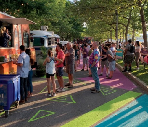 A lively outdoor scene with a crowd gathered around food trucks in a park, surrounded by trees and colorful pavement.