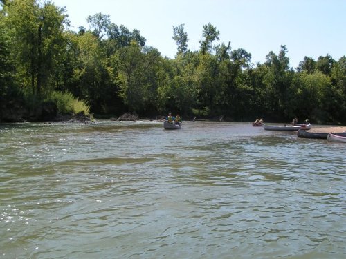 A serene river scene with canoes and people enjoying nature, surrounded by lush green trees under a clear blue sky.