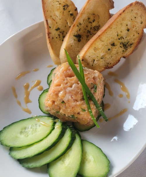 A plate featuring a salmon tartare with cucumber slices and toasted garlic bread.