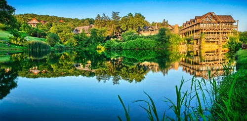 A serene lake reflects a rustic lodge surrounded by lush greenery and trees under a clear blue sky.