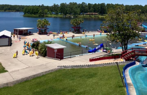 A sunny water park scene with people enjoying the pool, slides, and lake views surrounded by trees.