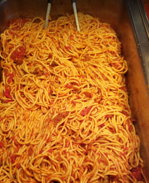 A large tray of spaghetti with a rich tomato sauce, served with tongs for serving.