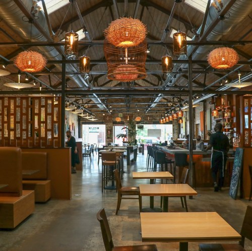 A spacious restaurant interior with wooden tables, woven light fixtures, and a bar area, featuring a warm, inviting atmosphere.