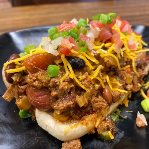 A loaded hot dog topped with chili, cheese, diced tomatoes, onions, and green onions on a black plate.