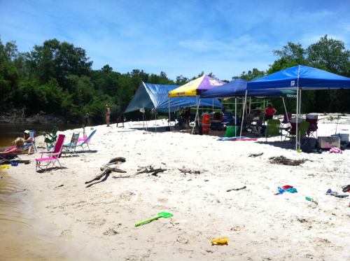 A sandy beach scene with colorful tents, chairs, and scattered toys, surrounded by trees and a calm river.