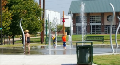 Children playing in a splash pad while an adult watches nearby, surrounded by greenery and a modern building.