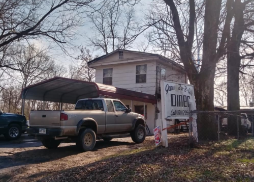 A two-story diner with a carport, surrounded by trees, featuring a sign that reads "Open Bar-B-Q Diner."