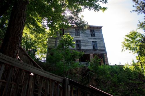 Abandoned wooden building surrounded by trees, with sunlight filtering through the leaves. Steps lead up to it.