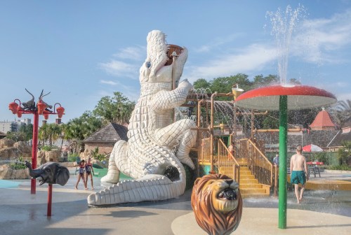 A water park featuring a large crocodile slide, animal sculptures, and children playing in the splash area.