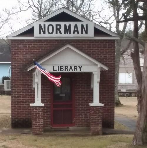 A small brick building with a sign reading "NORMAN" and "LIBRARY," featuring an American flag at the entrance.