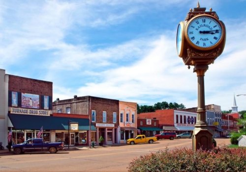 A charming small town street with a vintage clock, shops, and colorful vehicles under a blue sky.