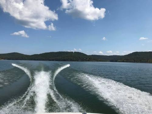 A scenic view of a lake with boat wake, surrounded by green hills and a clear blue sky with fluffy clouds.