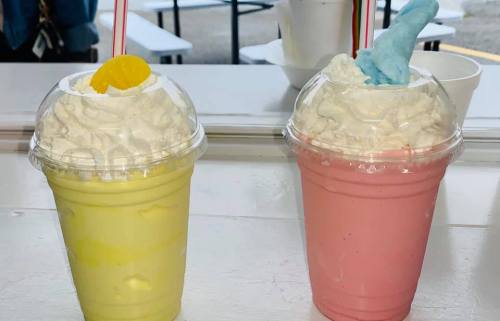 Two colorful milkshakes in clear cups, one yellow with a lemon slice and whipped cream, the other pink with blue cotton candy.