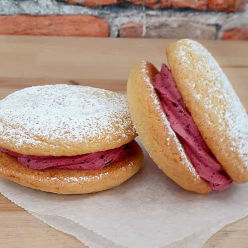 Two cookie sandwiches with pink filling, dusted with powdered sugar, on a wooden surface against a brick background.