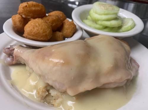 A plate featuring a chicken leg covered in creamy sauce, served with cornbread bites and cucumber slices.