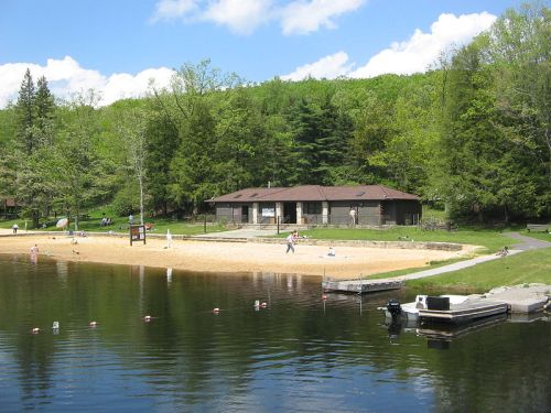 A serene lake scene featuring a sandy beach, a building, and lush green trees under a partly cloudy sky.