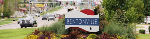 Sign welcoming visitors to Bentonville, surrounded by colorful flowers and greenery, with a busy road in the background.