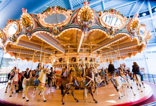 A colorful carousel with ornate decorations, featuring riders on horses and bright lights in a lively indoor setting.