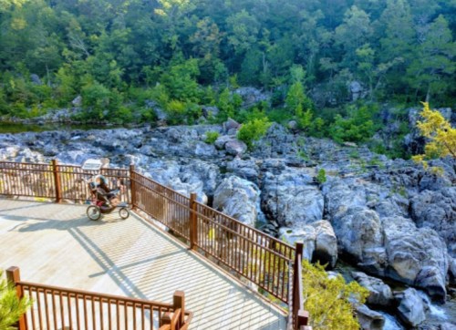A person with a stroller on a wooden deck overlooking rocky terrain and lush greenery.