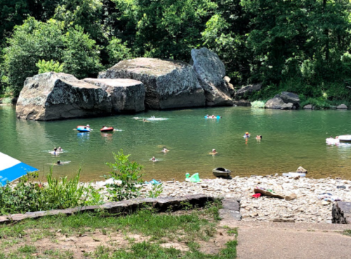 People swimming and relaxing in a river surrounded by large rocks and lush greenery on a sunny day.