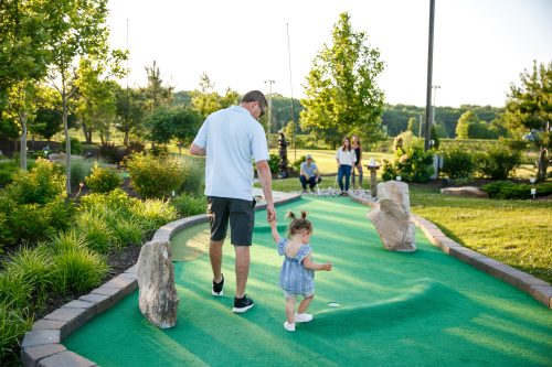 A man and a young girl walk along a mini-golf course, with others playing in the background on a sunny day.